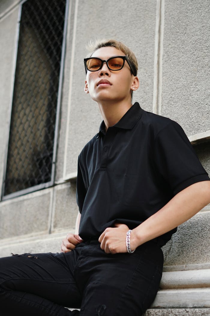 Stylish Asian man wearing a black polo and sunglasses sitting casually in an urban setting.