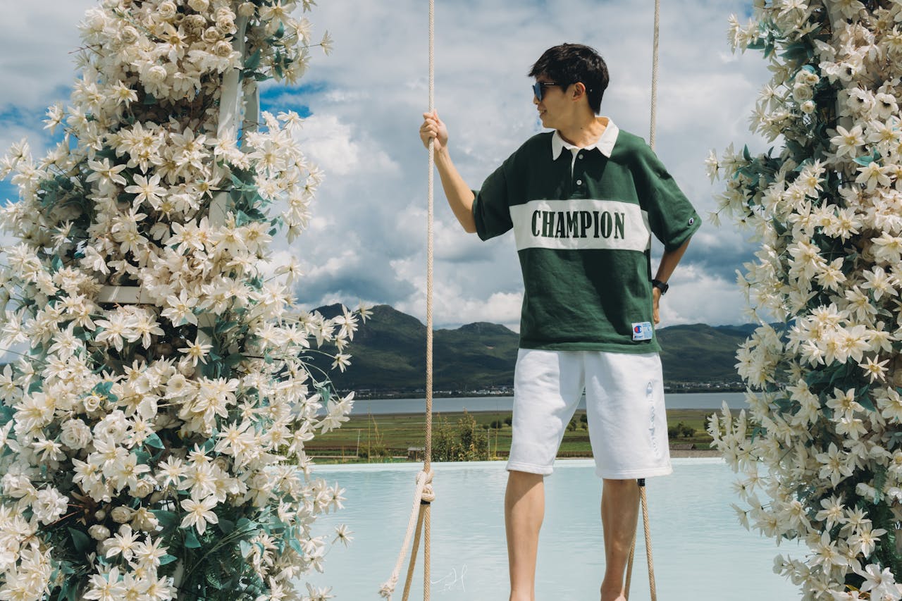Young man on a swing adorned with flowers, enjoying a summer day with scenic mountain views.
